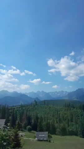 A video captures a scenic view from a vehicle traveling on a road. The foreground features trees and a glimpse of a building. The middle ground displays a dense forest and a small house. In the background, majestic mountains under a clear blue sky dotted with white clouds complete the picturesque landscape. The footage seems to be taken on a sunny day.