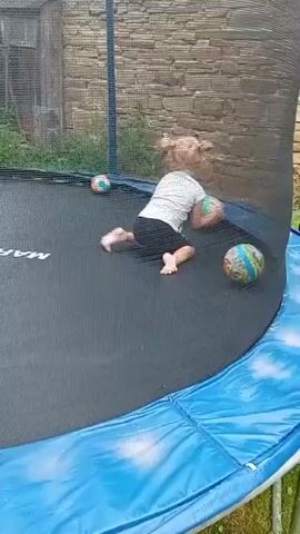 A young girl with blonde hair pulled back in a ponytail is playing on a trampoline. She is wearing a white t-shirt and dark shorts. Several colorful balls are scattered on the trampoline around her. The trampoline has a black jumping mat with the "MAXIMEX" logo visible, and a blue padded edge. The scene is outdoors, with a stone building and some greenery in the background. The lighting suggests it is daytime. The girl appears to be crawling and reaching for a ball, engaged in play.