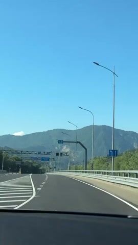 The video captures a view from inside a vehicle driving on a multi-lane highway. The road is clear and paved, with white dashed lines indicating lanes and solid white lines marking the edge. Overhead gantries display blue road signs with white directional arrows, indicating lanes for straight ahead and turns. A white barrier runs along the right side of the road, separating it from the surrounding greenery and trees. In the distance, a mountain range with lush green slopes is visible under a clear blue sky. Several modern streetlights are positioned along the roadside. The overall impression is that of a well-maintained highway in a scenic, mountainous region on a sunny day.