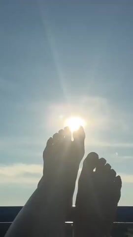 A person's bare feet are silhouetted against a bright sky. The sun is visible through the toes of the feet. The sky is blue with some scattered clouds. The image appears to be taken from a low angle, looking up at the feet and the sky.