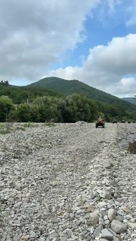 The video shows a rocky riverbed with a group of people riding ATVs in a line. The ATVs are yellow and driven by people wearing casual clothing. The riverbed is covered in gray and white stones, with some green trees and vegetation on either side. In the background, there are green hills and mountains under a cloudy blue sky. The ATVs are moving forward, kicking up dust from the rocky terrain. The scene appears to be an outdoor recreational activity in a natural landscape.