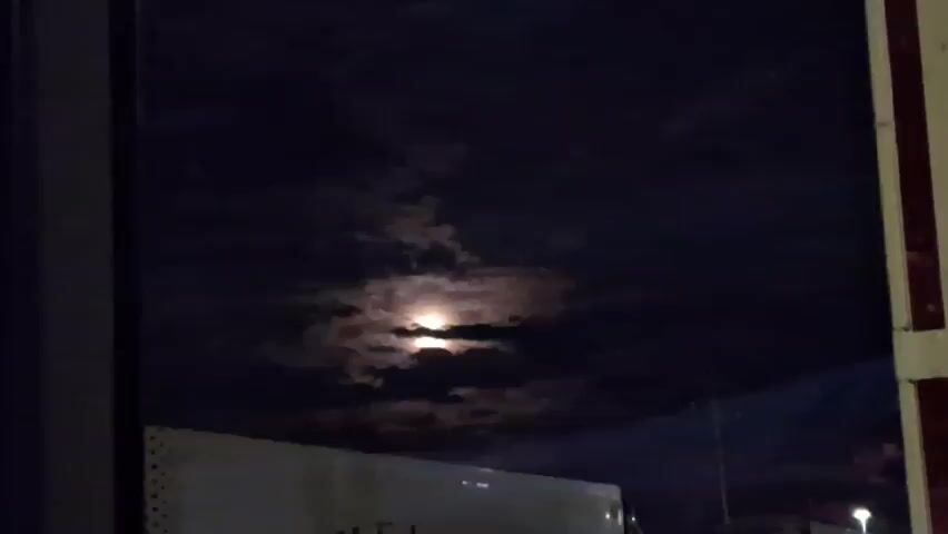 The video shows a nighttime scene. The moon is visible through scattered clouds, casting a glow on the sky. In the foreground, there is a reflection in what appears to be a window. The reflection shows parts of a white vehicle, possibly a truck, and dark horizontal lines on the right side of the frame, which might be part of a building's structure or a vehicle. The overall lighting is dim, indicating nighttime.