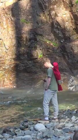 A person stands near a waterfall, looking at their phone. The person is wearing a green t-shirt, light-colored pants, and sneakers, with a red bag slung over their shoulder. The waterfall cascades down a rocky cliff face, and a rainbow is visible in the mist. The ground around the waterfall is covered in rocks.