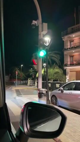 A night-time point-of-view video from a car shows the car turning right at an intersection. A traffic light changes from green to red. Palm trees line the street, illuminated by streetlights. There are several waste bins on the side of the road and a white building with balconies. The car's side mirror is visible in the foreground.