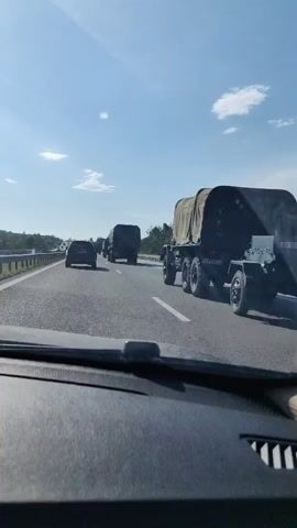 The video shows a view from inside a car, driving on a highway on a sunny day with a few clouds in the sky.  Multiple military trucks are in front of the car, traveling in the same direction. The trucks are covered with tarps and have visible license plates. A dark colored civilian vehicle is also in front of the car. Trees line the side of the highway.