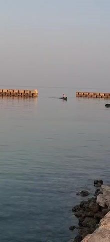 A person is paddling a small boat on calm water. The boat is green and white, and the person is wearing a white shirt and red shorts. The person is facing away from the camera, towards a breakwater. The breakwater is made of concrete blocks and stretches across the water. The water is clear and reflects the sky. The sky is a pale blue, and the sun is setting. The overall mood of the video is peaceful and serene.