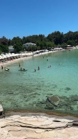 The video shows a clear, turquoise bay with several people swimming and relaxing on a sandy beach. The beach is lined with trees and lounge chairs. There are also a few boats anchored in the bay, including a small red rowboat and two sailboats. The water is very clear, and you can see the rocky bottom. The sky is bright blue and clear.