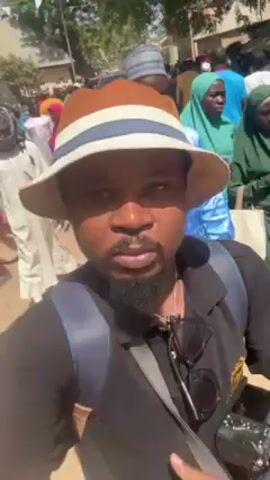 The video shows a man taking a selfie in a crowded outdoor setting. He is wearing a light brown hat with blue and white stripes, a black shirt, and a backpack. Sunglasses are hanging on his shirt. A digital camera is visible in his right hand. The background is filled with people, most wearing traditional attire, with a variety of colors, including green, pink, yellow, and blue. The setting appears to be a street or open area in a town or village, with buildings and trees visible in the background. The lighting is bright, suggesting a sunny day. The camera is shaky, indicating the person is walking through the crowd.