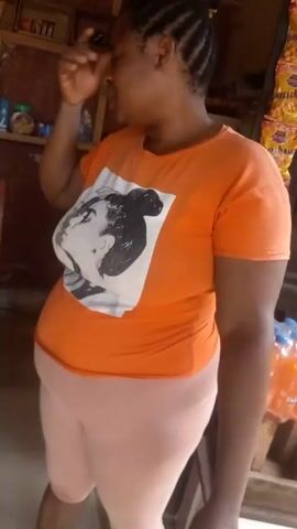 A person with braided hair, wearing an orange shirt with a graphic print and light pink pants, stands in what appears to be a small shop or store. Shelves are filled with various products, including bottles of Fanta, chips, and other grocery items. The person is seen adjusting their shirt and briefly looking at the camera before reaching towards the shelves. The overall setting seems to be a modest retail environment.