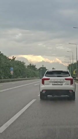 The video shows a white Haval SUV driving on a highway. The vehicle is in the right lane, moving away from the camera. The sky is cloudy with some sunlight breaking through. There are trees and roadside lights visible on the left side of the highway. Another car is visible in the distance in the left lane. The license plate of the Haval is partially visible and appears to be from Russia. The interior of the car from which the video is being filmed is not visible. The video duration is approximately 5 seconds.