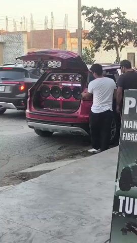 In a wide shot taken at what appears to be a street in Trujillo, Peru, a red SUV with an extensively modified sound system in its open trunk is parked on the side of a road. The trunk is packed with multiple large speakers, subwoofers, and illuminated panels. Additional spotlights or speakers are mounted on the vehicle's roof rack.

Two men are visible near the car. One man, wearing a white t-shirt and dark pants, is bent over, facing into the open trunk, seemingly adjusting or working on the sound equipment. Another man in a dark t-shirt is partially visible to the right, also near the car.

In the background, another car drives past, and residential or commercial buildings are visible. A large, dark trash bin stands further down the street. The lighting suggests it is either early morning or late afternoon. A sign on the right side of the frame is partially visible, showing text fragments like "P," "NA," "FIBR," and "TU."