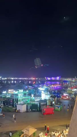 The video opens with a dark night sky above a bustling waterfront scene. A large, illuminated drone formation resembling a sailing ship with intricate light patterns is visible in the sky. Below, a vibrant harbor is filled with boats, many of which are also lit up. The waterfront promenade is a hive of activity with numerous small kiosks and structures adorned with colorful lights. People are seen walking around and congregating in various areas. The overall atmosphere is festive and lively, suggesting an event or a popular night-time destination. The camera is positioned at an elevated viewpoint, offering a wide panorama of the illuminated harbor and the drone display.