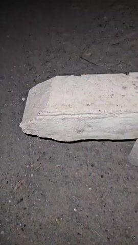 The video captures a close-up, horizontal pan across a concrete structure in a dark, possibly nighttime, environment. A long, rectangular, light-colored concrete slab or beam occupies the center of the frame, resting on several smaller, irregularly shaped concrete blocks. The ground beneath is dark, rough, and uneven, resembling dirt or gravel, with some small debris visible. The camera slowly tracks from left to right, maintaining focus on the concrete objects and the ground directly below them. Both the slab and the supporting blocks exhibit a coarse, weathered texture. The low lighting conditions, likely from an artificial source such as a flashlight, illuminate only the immediate foreground. There are no people, animals, or specific activities taking place. The scene appears to be an outdoor setting, potentially a construction site or an undeveloped area, located in Cam Đức, Vietnam.