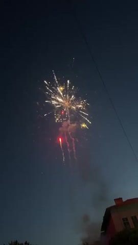The video is filmed vertically and shows fireworks lighting up the night sky against a backdrop of a dark blue sky with faint streaks of light blue. In the bottom right corner, a terracotta-colored building with a dark roof and a small chimney is visible. Streaks of light from the fireworks are seen ascending from the bottom of the frame. One red firework bursts into a shower of white and red sparks, followed by a yellow firework exploding with white and yellow sparks. Smoke rises into the sky, obscuring parts of the display. Throughout the video, more fireworks ascend and explode in various patterns of white and yellow sparks, creating a continuous display of light and color against the night sky. The video captures the fleeting beauty of fireworks, highlighting the contrast between the dark sky and the bright bursts of light.