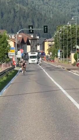 A cyclist is riding down a paved road in a town, with buildings and trees lining the street. A white camper van is parked on the side of the road, and several people are walking on the sidewalk. In the background, a dense forest covers a mountain. The sky is clear and blue. A traffic light is visible, currently showing green.