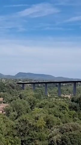 This video shows a long, elevated bridge with multiple supports crossing over a dense, green forest. The sky is blue with scattered clouds. The bridge appears to be made of concrete and is a uniform dark gray color. The trees are lush and varied in texture, suggesting a natural, undeveloped area. In the distance, a mountain range is visible under the horizon. There are no people or vehicles clearly visible on the bridge or in the immediate vicinity. The camera seems to be positioned at a distance, looking towards the bridge from a higher vantage point.