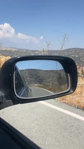 The video shows a view from a car driving on a road. The perspective is from the passenger side, looking out the side mirror. The side mirror reflects a winding road that goes through a hilly, dry, and mountainous landscape. The sky is blue with some white clouds. There are sparse trees and vegetation visible along the roadside and in the distance. The road has a white line marking the edge.