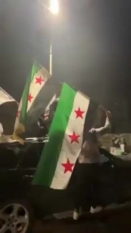 At night, a crowd of people stand on and around a black car, waving flags and cheering. The flags are predominantly green, white, and black with red stars; they are likely the flags of Syria. Some individuals are holding white flags with Arabic text. A street light is visible in the background. People are wearing a variety of clothing, and some are wearing head coverings. Several people are holding up their phones, presumably to record the event. The overall atmosphere suggests a celebration or protest.