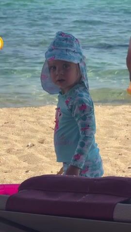 A young child wearing a sun hat and a long-sleeved swimsuit with a flamingo pattern stands on a sandy beach. The child looks towards the camera, then smiles and claps their hands. In the background, the ocean is visible, and another person, partially obscured, is standing in the water. A beach chair is in the foreground.