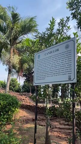 A sign stands in front of a weathered brick structure, possibly a ruin, surrounded by lush green foliage and palm trees. The sky above is clear and blue. The sign appears to be informational, with text and an emblem. The overall scene suggests an outdoor historical or archaeological site.