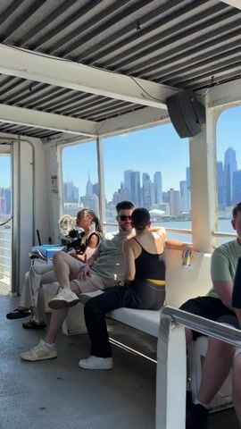 The video shows the interior of a ferry with people seated on a bench. The ferry has large windows offering a view of a city skyline and water. Sunlight streams in through the windows, and the ceiling is lined with metal slats. On the bench, three people are visible from the waist down. Two individuals, a man and a woman, are seated next to each other, with the man on the left. The woman on the right has dark hair and is wearing a black tank top with a yellow stripe. The man has dark hair and a beard and is wearing a light-colored t-shirt and dark pants. To the left of the couple, a woman with braided hair is seated, wearing a light-colored outfit. She is holding a small dog. Her legs are crossed. Her shoes are white sneakers. The man's shoes are white sneakers. The woman's shoes are dark slides. A man in a blue uniform, possibly a crew member, is standing in an open doorway to the left of the ferry passengers. He is wearing a blue shirt and black pants. He is interacting with a woman who is partially visible in the doorway. The man in uniform is wearing black gloves. There is a blue cooler or storage bin behind the seated passengers.
