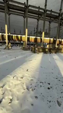 The video shows the interior of a large, cylindrical structure, possibly a cooling tower. The ground is covered in snow and debris. A series of metal support beams and large pipes run horizontally across the interior. The camera tilts upward to reveal the open top of the structure, through which the sky is visible. The sky is blue with some clouds. The structure is made of concrete and appears to be in a state of disrepair. No people are visible.