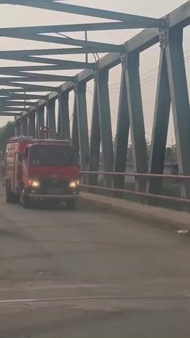 A red fire truck with flashing red lights drives across a bridge. The truck has the word "Firefighter" written on its side in white letters. Two people on motorcycles follow behind the fire truck. The bridge is a truss bridge with dark metal beams. The sky is overcast.