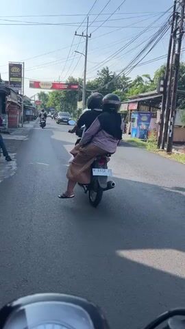 The video captures a street scene with several motorcycles. In the foreground, a portion of a motorcycle handlebar and dashboard is visible. A person wearing a helmet and a dark jacket is on a motorcycle to the left, parked and facing away from the camera. Further down the street, a motorcycle with two passengers, one in front and one behind, is moving away from the camera. The passenger in the rear is wearing a headscarf and a patterned skirt. Several other motorcycles are visible further down the road, also moving away. The environment appears to be a paved street lined with buildings, possibly shops, with power lines overhead. A white car is parked on the right side of the street. The sky is bright and sunny.