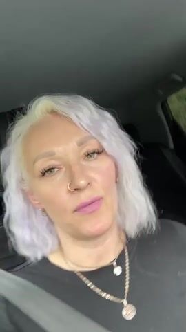 A medium shot shows a woman with short, light purple hair and fair skin sitting in a car. She is wearing a black top and a seatbelt. She has on makeup including pink lipstick and mascara. She is wearing multiple necklaces and earrings, and has a nose ring. Her fingernails are painted red. The woman brings her hand up to her face and covers her mouth with her fingers. She appears to be talking or singing. The background is dark and blurry, suggesting the interior of a vehicle.