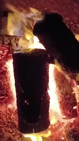 The video shows a close-up view of logs burning in a fireplace. The logs are arranged vertically and horizontally, with flames licking around them. The fire is a mix of orange, yellow, and blue, indicating intense heat. The surrounding embers are a bright red, suggesting that the fire is well-established. The scene is dimly lit, with the fire being the primary source of light. The video appears to be shot with a handheld camera, as there is some slight movement and shakiness. The audio, if present, is not described.