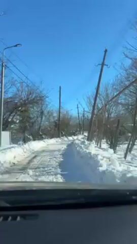 The view from inside a car shows a snow-covered road, bordered by snowbanks, flanked by power lines and bare trees under a clear blue sky. The car is moving forward.