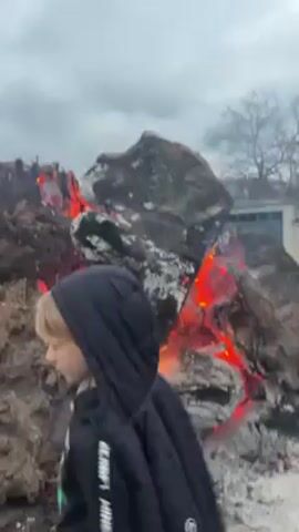 The video shows a young person with blonde hair wearing a black hoodie walking in front of a pile of burning debris. The debris appears to be composed of charred wood and other materials, with visible flames and glowing embers. The background is overcast, with a glimpse of a building in the distance. The camera movement is unsteady, suggesting handheld recording.