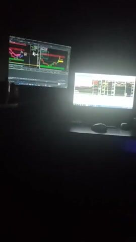 The video shows two computer monitors in a dark room. The monitor on the left displays a trading chart with red and green candlesticks. The monitor on the right also displays charts and financial data. There are two computer mice visible on the desk in front of the monitors. The room is dimly lit, with the glow of the monitors being the primary light source. No people are visible in the video.