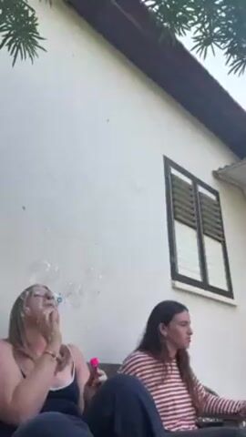 Two young women are seated outside a white building. The woman on the left is blowing bubbles with a pink wand. The woman on the right is watching the bubbles. The building has a dark brown trim and a window with closed shutters. Green foliage is visible above the building. The woman on the left is wearing a black tank top and glasses. The woman on the right is wearing a red and white striped shirt and dark pants.