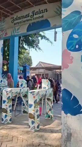 The video shows an outdoor scene, likely an entrance or exit gate, decorated with blue and white painted foliage. Above the gate, there is a sign that reads "SELAMAT JALAN Sampai bertemu kembali," which translates to "Have a good trip, see you again." Several people are present, some of whom are using turnstiles. Two young males wearing red jackets with text on the back are walking towards the camera and then past the turnstiles. Other individuals, including women wearing hijabs, are either waiting to pass through the turnstiles or have already done so. In the background, there are buildings and more people. The environment appears to be sunny with clear skies.