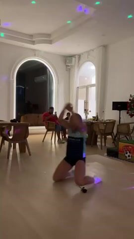 A young blonde woman with a teal and black athletic top and black shorts is performing a dance routine on her knees on a wooden floor in a brightly lit room. She is looking upwards with her arms outstretched. There are colored lights projected onto the ceiling and floor. In the background, there are several wooden tables and chairs, an arched window, and two men seated at a table in the distance. A red rose lies on the floor near the woman.