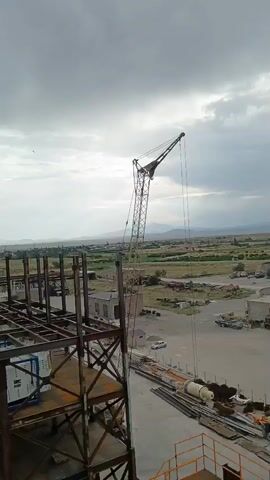 The video shows a construction site with a tall crane positioned to the right of the frame. In the foreground, a partially constructed steel building is visible with beams and columns forming its framework. Below the steel structure, a white piece of equipment with blue accents is partially visible. In the background, a rural landscape with scattered buildings and distant mountains can be seen under a cloudy sky. A white car is parked near a building in the mid-ground. The crane's boom extends upwards and to the right, with a cable hanging down. The scene appears to be an industrial or construction area.