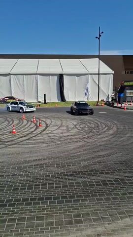 Two cars are drifting in a wide paved area with scattered orange cones. The car on the left is white and appears to have its hood open. The car on the right is dark purple and has a spoiler. Both cars are producing smoke from their tires as they drift in opposite directions, leaving tire marks on the pavement. In the background, there is a large white tent, a dark brown building, an inflatable archway, and several people standing around. The sky is clear and blue.