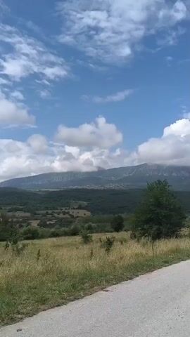 This video captures a scenic view of a rural landscape in Ruska Bela, Bulgaria. The camera pans across a rolling terrain with dry, golden grass in the foreground, suggesting a warm, possibly late summer or early autumn day. A paved road curves into the frame from the right, indicating a journey or a passing view.

Beyond the field, a dense, dark green forest stretches across the middle ground, punctuated by scattered trees. In the distance, a mountain range rises, its peaks partially obscured by dramatic, white cumulus clouds that drift across a bright blue sky. A small village or settlement is visible nestled on the slopes of the mountains, adding a touch of human presence to the vast natural setting.

The overall atmosphere is one of tranquility and natural beauty. The weather appears to be pleasant, with a mix of sun and clouds creating dynamic lighting. There is no visible text in the video. The camera's perspective suggests it might be mounted on a moving vehicle, providing a dynamic viewpoint of the Bulgarian countryside.