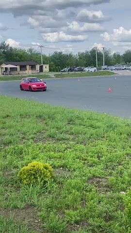 This is a short video capturing a driving event at Svetloe Pole, Russia. The primary subjects are two cars: a red Volkswagen Golf GTI and a red convertible sports car, possibly a Mazda MX-5 Miata. The Golf is in the foreground, being driven on a paved track with a grassy area beside it. The sports car is further back on the track, also in motion. Several orange traffic cones are visible on the track, indicating a course or training exercise.

The scene is outdoors on a track that appears to be a dedicated facility for automotive activities. In the background, there are trees, a building with white siding, and more parked cars. Beyond the track, a wide expanse of green fields and distant trees suggests a rural or semi-rural setting. The sky is cloudy, but the lighting suggests it is daytime, likely afternoon.

The activity captured is the movement of these cars on the track, suggesting a driving session, possibly a track day, autocross event, or driver training. The cars are shown from various angles as they navigate the course. There are no people clearly visible within the vehicles, but the presence of the cars and cones implies human activity and organized event.

Notable details include the bright red color of both cars, contrasting with the green of the grass and the gray of the asphalt. The black rims on the Golf are a distinctive feature. The weather appears overcast, but not rainy. The location cues, such as the landscape and a distant industrial-looking building, are consistent with an outdoor driving facility in Russia. No visible text is present in the video. The overall mood is energetic and focused on automotive performance.