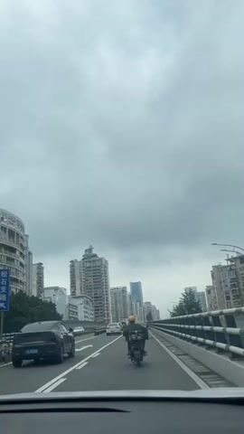 The video shows a view from a vehicle traveling on a multi-lane highway or elevated road. The sky is overcast with heavy gray clouds. On the left side of the road, there are several tall buildings, some appearing to be residential complexes with balconies, and others are modern skyscrapers. On the right side of the road, there is a barrier with white and green posts. The road itself has multiple lanes separated by white lines. In the foreground, the dashboard or interior of the vehicle from which the video is recorded is visible. Several cars are on the road, including a black sedan in the left lane and a white car further ahead. A person on a scooter wearing a yellow helmet is in the right lane, moving in the same direction as the camera vehicle. Traffic appears to be moderate, with cars and the scooter moving along the road. The overall impression is an urban environment on a cloudy day.