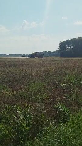 A wide shot shows a large agricultural machine with a yellow tank on the back driving across a dry field under a clear blue sky. The machine is kicking up dust as it moves. Trees line the horizon in the background. The foreground is filled with tall, green grass.