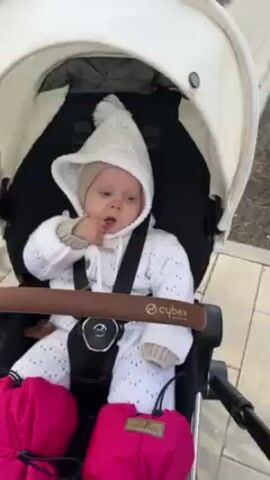 A baby is seated in a Cybex stroller on a paved surface. The baby is wearing a white knitted outfit including a hat with a pointed top. The baby has their finger in their mouth and is looking forward. Two bright pink bags labeled "Little Baby" are attached to the front of the stroller. The stroller has a white canopy and a brown handlebar. A sidewalk and some sparse vegetation are visible in the background.