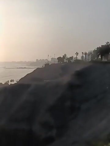 The video shows a coastal view at sunrise or sunset, with the sun reflecting on the water. The shot starts with a view partially obscured by a dark, rocky landmass in the foreground. As the camera moves, the scene reveals the ocean, a beach, and a city skyline in the distance, all hazy due to the sun’s brightness. Waves can be seen gently rolling towards the shore. The coastline extends along the horizon, and the camera continues to shift, revealing a grassy edge of a cliff.