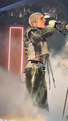 The video shows a bald, tattooed man performing on stage, singing into a microphone. He is wearing a unique, shimmering, olive-green jumpsuit with a structured jacket. The jacket has exaggerated, layered shoulders and a shiny, metallic finish. He is also wearing a belt with large, silver embellishments. Smoke or fog is visible around his feet, and stage lighting illuminates the scene from behind. The man's arms are raised, and he appears to be in the midst of an energetic performance.