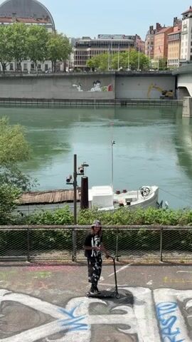 The video opens with a wide shot of an urban riverside scene, likely in Lyon, France. In the foreground, a young person with long dark hair, wearing a patterned beanie, black t-shirt, and black and white patterned pants, stands on a paved surface covered in graffiti. They hold a black kick scooter, facing the camera with a slight smile. A metal fence separates them from a wide, calm river with greenish-blue water.

On the river, a white boat with a red flag is docked, with some utility poles nearby. The opposite bank features a substantial concrete retaining wall, topped by a road or bridge. A large mural is painted on this concrete wall.

The video then zooms in to reveal the mural in detail. It depicts a boy with messy dark hair, round glasses, and headphones, intently reading a book. The boy is rendered in a black and white, graphic style. The book he holds is open, revealing a vibrant, colorful illustration featuring a bright yellow background and a green, abstract figure, strongly resembling the artwork of Keith Haring. The artist's name, "Haring," is visible on the red spine/cover of the depicted book. To the left of the reading boy, another smaller, partially visible figure is lying down, also in the same black and white style.

Above the wall, an urban landscape unfolds with multi-story buildings, green trees, and glimpses of a road or bridge structure. A yellow excavator is visible on the far right of the opposite bank. The scene is bathed in bright daylight under a clear sky, suggesting a sunny day.