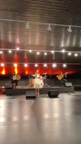 A wide shot shows a band performing on a stage in a large empty hall. The band consists of a female vocalist, a guitarist, an accordion player, and a drummer. The female vocalist stands center stage wearing a white dress and blue shoes. She is holding a microphone and singing. The guitarist is seated to her right, wearing a black shirt and pants. The accordion player is seated to her left, also wearing a black shirt and pants. The drummer is in the back center of the stage behind the vocalist. Behind the band are red stage lights arranged in a line. String lights are also visible above the stage. The floor of the hall is shiny and reflects the lights. The ceiling of the hall is made of metal beams.