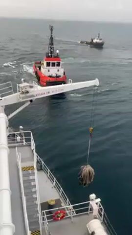 The video shows two boats on the ocean, as viewed from the deck of a larger vessel. In the foreground, a crane is lifting a mass of rope, seemingly transferring it from the deck. A person can be seen standing on the deck, observing the operation. A red and white tugboat is prominently featured in the middle ground, moving through the water. Further in the distance, a second, smaller boat is visible, also moving on the water. The sky is overcast, contributing to the overall dull lighting of the scene.