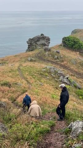 The video shows three people hiking down a grassy, rocky trail near the ocean. One person, dressed in black, stands still while the other two, dressed in a blue jacket and a tan jacket, walk ahead down the path. The camera pans to reveal a wider view of the coastline, showing a small beach and a bay. The sky is overcast and the water is calm. The landscape is covered in low-lying vegetation and scattered rocks.