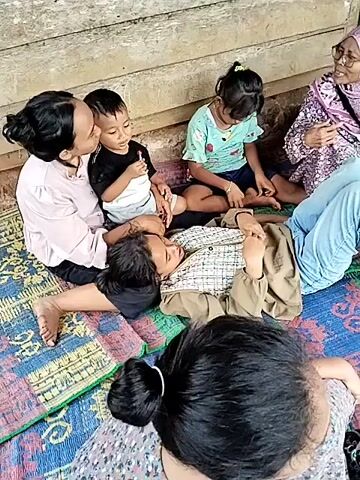 A group of six people, including three children and three adults, are sitting on mats on the ground in front of a wooden structure. One adult woman, wearing a pink shirt and glasses, is speaking to another adult woman wearing a light purple headscarf and a patterned shirt. A young boy is sitting between two adult women, one of whom is wearing a pink shirt, and is being kissed on the cheek by the woman in the pink shirt. Another young child, a girl, is sitting on the mat with her legs crossed. Two other adult women are lying down on the mats, with one of them resting her head on the lap of another. The background appears to be a wall or structure made of weathered wood planks.